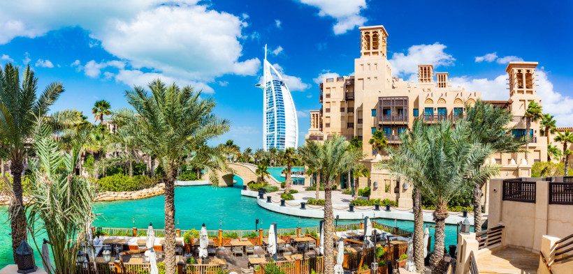 Dubai Panoramaaufnahme eines luxuriösen Resorts in Dubai mit Wasserkanälen, Palmen und traditionellen Gebäuden im arabischen Stil. Im Hintergrund ragt das berühmte Hotel Burj Al Arab mit seiner segelförmigen Architektur in den Himmel. Im Vordergrund befinden si