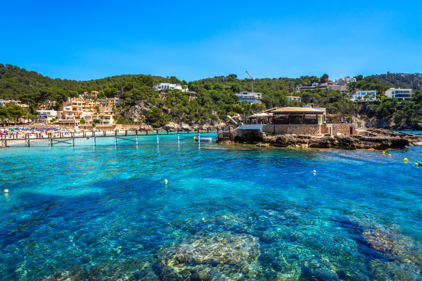 Camp de Mar mit Inselrestaurant La Illeta und klarem Wasser Blick auf das Restaurant La Illeta in Camp de Mar, erreichbar über einen Holzsteg, mit kristallklarem Wasser und bewaldeten Hügeln im Hintergrund.