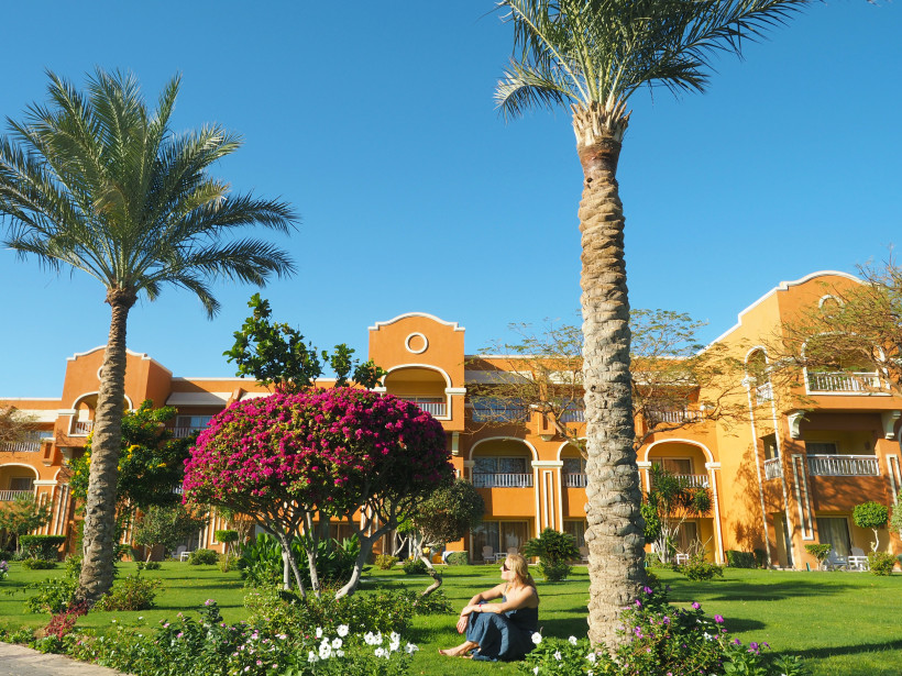 Frau sitzt im gepflegten Garten einer farbenfrohen Hotelanlage mit Palmen, Blumen und blauem Himmel.