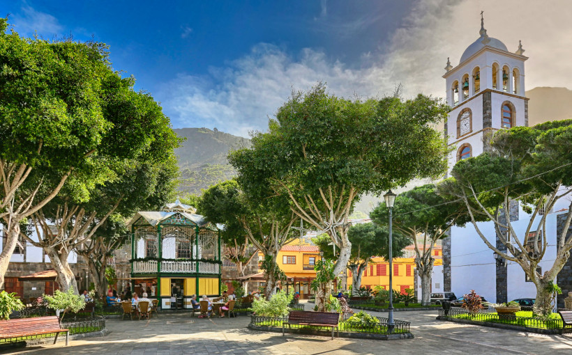Idyllischer Dorfplatz in Garachico auf Teneriffa mit Schatten spendenden Bäumen und Kirchturm Ruhiger Dorfplatz in Garachico auf Teneriffa mit großen Schattenbäumen, Sitzbänken, einem historischen Musikpavillon und dem weißen Kirchturm der Iglesia de Santa Ana im Hintergrund, eingerahmt von Bergen und blauem Himmel.