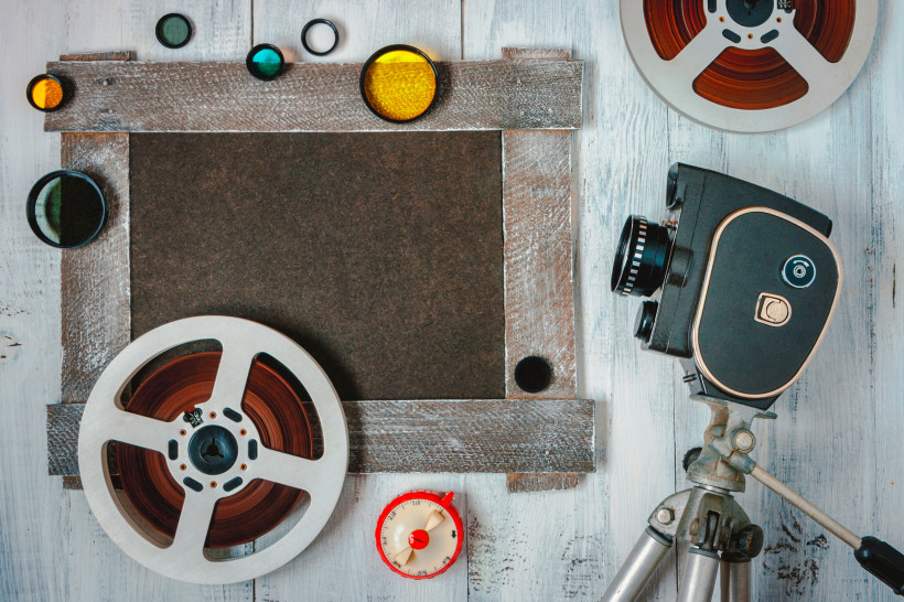 Ägypten - Sharm el Sheikh Vintage-Filmausrüstung auf hellem Holzhintergrund: Eine klassische Filmkamera auf einem Stativ rechts, Filmrollen mit braunem Band links, Farbfilterlinsen in Gelb, Grün und Blau sind dekorativ rund um einen schwarzen Rahmen angeordnet.