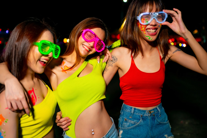 Thailand - Koh Samui Das Bild zeigt drei junge Frauen, die nachts bei einer Party ausgelassen feiern. Sie tragen auffällige Neonbrillen in Grün, Pink und Blau, dazu bunte Tops – zwei in Neongelb und Rot, eine in Jeansshorts. Ihre Gesichter und Körper sind mit fluoreszierender
