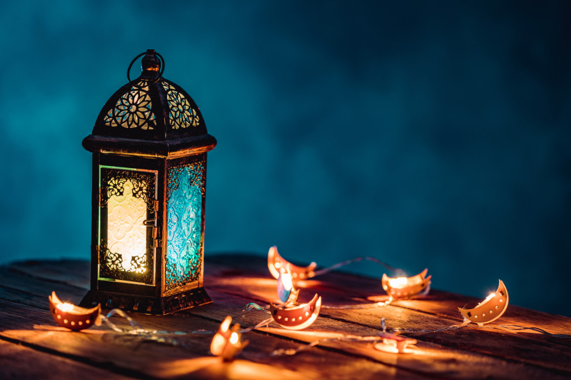 Ras al Khaimah, Ramadan Eine orientalische Laterne mit buntem Glas leuchtet auf einem Holztisch, umgeben von dekorativen Lichterketten mit Mond- und Sternmotiven