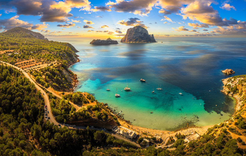 Ibiza - Es Vedrà Blick von der Küste bei Cala d’Hort auf die unbewohnte Felseninsel Es Vedrà vor Ibiza. Türkisfarbenes Meer, mehrere Segelboote im Wasser und ein strahlender Himmel mit Wolken bei Sonnenuntergang.