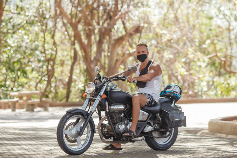 Thailand - Phuket Ein Mann mit kurzer Frisur sitzt auf einem schwarzen Motorrad. Er trägt ein weißes Tanktop, Shorts, schwarze Sandalen und eine schwarze Gesichtsmaske. Neben ihm auf dem Sitz liegt ein Helm. Im Hintergrund sind Bäume mit hellem Sonnenlicht zu sehen.