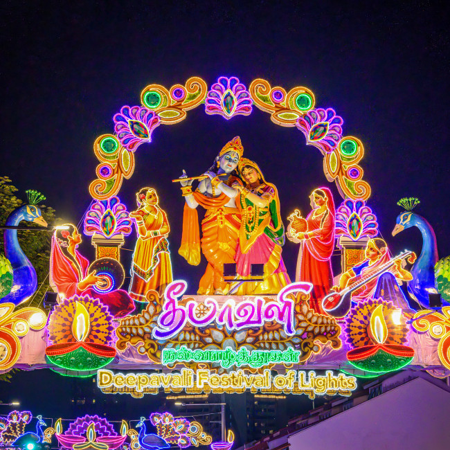 Singapur Farbenprächtiger Lichterbogen zum Deepavali Festival of Lights mit hinduistischen Gottheiten und Figuren. In der Mitte stehen Krishna und Radha, umgeben von weiteren Figuren, Pfauen und einem Lampensymbol. Die Szenerie ist in Neonfarben beleuchtet und det