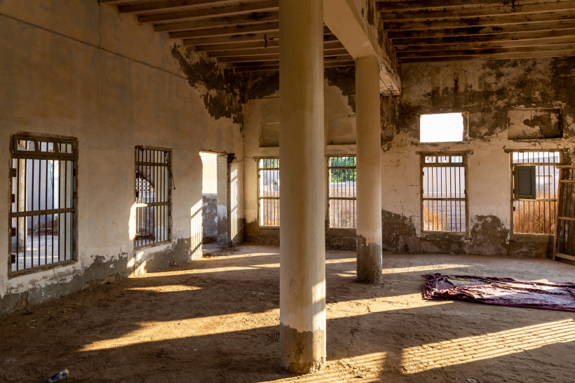 Innenansicht eines verlassenen Gebäudes in Al Jazirah Al Hamra mit Sonnenlicht, vergitterten Fenstern und historischen Mauern.