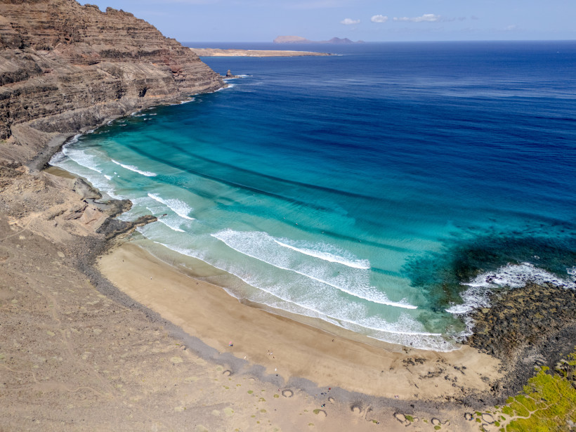 Der Playa de la Cantería nahe Órzola auf Lanzarote mit goldgelbem Sand, kräftigen Wellen und der imposanten Risco-Felskulisse – einer der wildesten Strände im Norden der Insel.