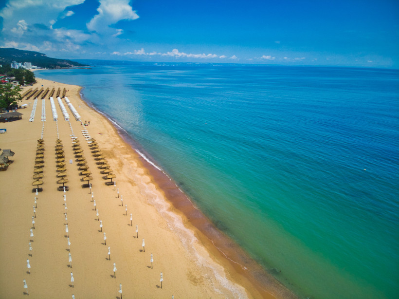 Goldstrand bei Tageslicht mit klarblauem Wasser und langen Reihen aus Sonnenliegen und Strohdach-Schirmen. Der weiche Sandstrand ist weitläufig, das Meer geht flach ins Wasser über. Am Horizont sind Hotels und bewaldete Hügel zu erkennen.
