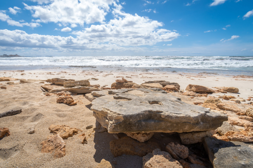 Es Trenc-Strand, Mallorca Felsiger Sandstrand mit flachen Steinen im Vordergrund und Wellen im Hintergrund unter teils bewölktem Himmel