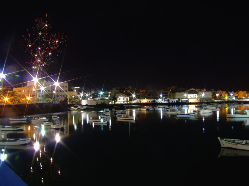 Lanzarote Nächtlicher Hafen mit kleinen Booten, beleuchtet von Laternen. Im Hintergrund erhellen bunte Feuerwerkskörper den Himmel über den Häusern der Stadt.