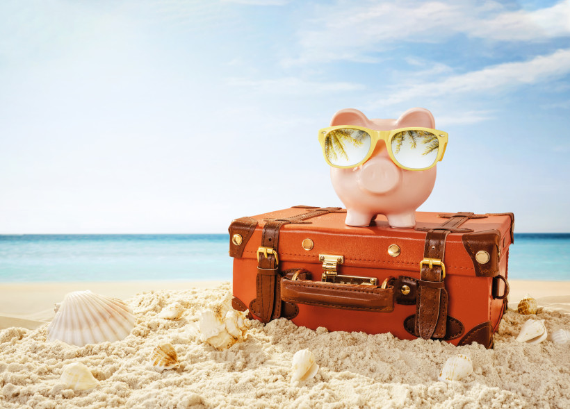 Sparschwein mit Sonnenbrille steht auf einem kleinen braunen Vintage-Koffer im Sand am Strand mit Muscheln und blauem Meer im Hintergrund