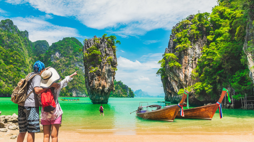 Paar an der Phang Nga Bay – Longtailboote und Felsenlandschaft in Thailand Paar steht am Strand der Phang Nga Bay in Thailand und zeigt auf die markanten Kalksteinfelsen, während Longtailboote am Ufer liegen.
