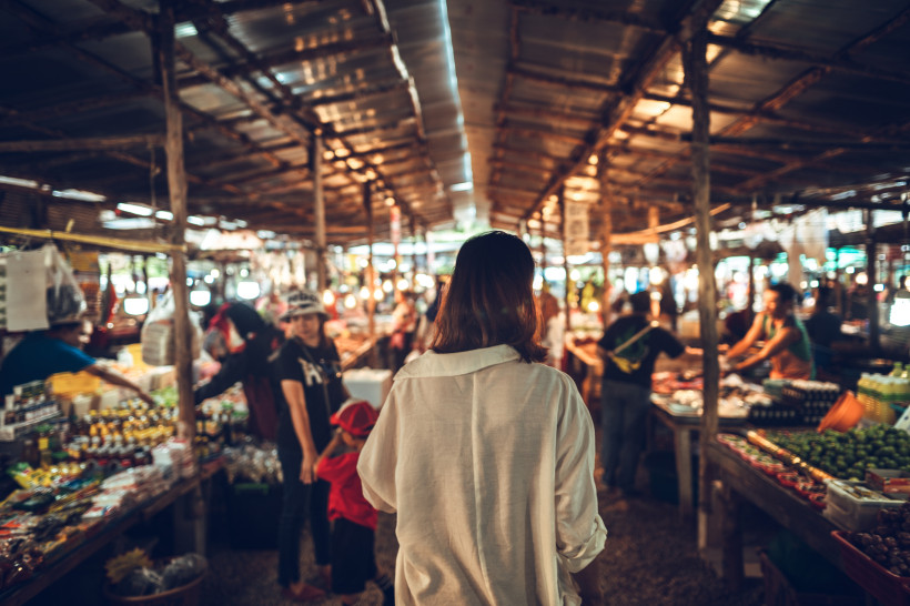 Thailand - Krabi Das Bild zeigt eine Frau mit schulterlangem Haar und weißem Hemd von hinten, wie sie durch einen traditionellen Markt in Südostasien läuft. Der Markt ist überdacht mit Wellblechdächern und von warmem Licht durchzogen. Links und rechts reihen sich Verkaufs