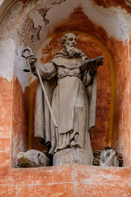 Mallorca Steinerne Statue eines Heiligen in einer Wandnische mit rot-orange Patina. Die Figur trägt ein langes Gewand, hält in einer Hand einen Stab mit einer Glocke und in der anderen ein aufgeschlagenes Buch. Zu Füßen der Statue liegt ein kleines steinernes Schw