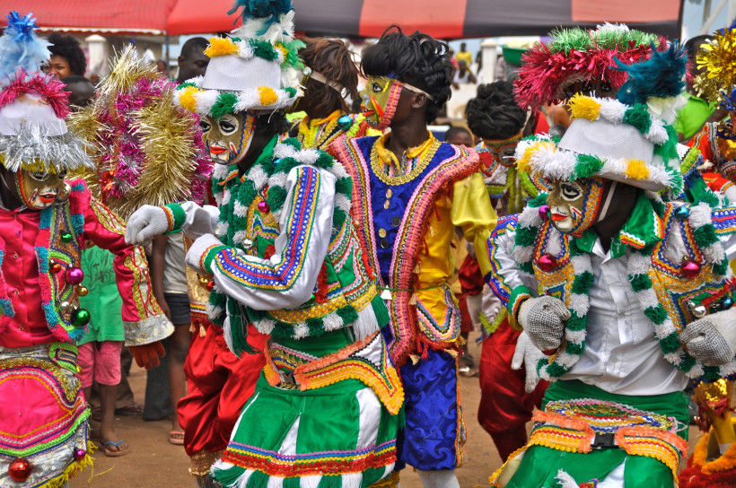Teilnehmende eines Straßenkarnevals in bunten, aufwendig verzierten Kostümen mit Masken, Pailletten und Federn. Die Personen tanzen in einer fröhlichen Parade auf einer Straße, umgeben von Zuschauer:innen. Die Szene wirkt lebendig und farbenfroh.