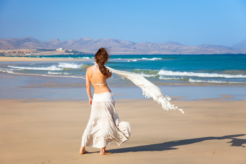 Frau in weißem Sommeroutfit steht am Strand von Fuerteventura und blickt auf das türkisfarbene Meer