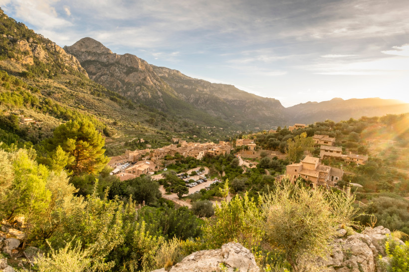 Mallorca - Fornalutx Eine weite Landschaft Mallorcas mit terrassenförmig angelegten Feldern, die sich an die Hügel schmiegen. Die Szenerie wird von kleinen, sandfarbenen Steinhäusern dominiert, die sich harmonisch in die Landschaft einfügen. Im Hintergrund erheben sich bewald