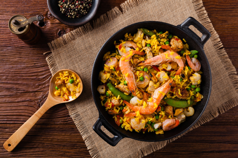 Paella in einer schwarzen Pfanne mit gelbem Reis, Garnelen, Meeresfrüchten und Gemüse auf einem Holztisch