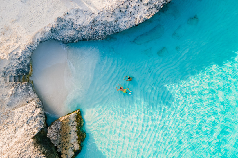 Zwei Menschen schwimmen in glasklarem, türkisblauem Wasser einer Bucht auf Aruba. Umgeben von hellen Felsen und Sand, eingefangen aus der Vogelperspektive.