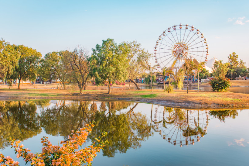 Volksfest in Aguascalientes Ein malerischer Park mit einem ruhigen See, in dem sich Bäume und ein großes Riesenrad spiegeln, umgeben von grüner Natur und einer warmen Abendstimmung