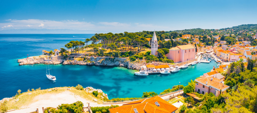 Kroatien Panoramablick auf einen malerischen Küstenort in Kroatien mit türkisblauem Wasser, kleinen Jachten im Hafen, einer großen Kirche mit Turm, roten Ziegeldächern und grüner Vegetation entlang der Küste.