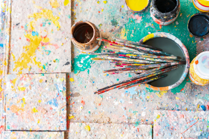 Ägypten - Marsa Alam Mehrere bunte, gebrauchte Pinsel stehen in einem Farbtopf auf einem mit Farbspritzern bedeckten Boden. Umgeben sind sie von offenen Farbdosen in kräftigen Farben wie Gelb und Schwarz.