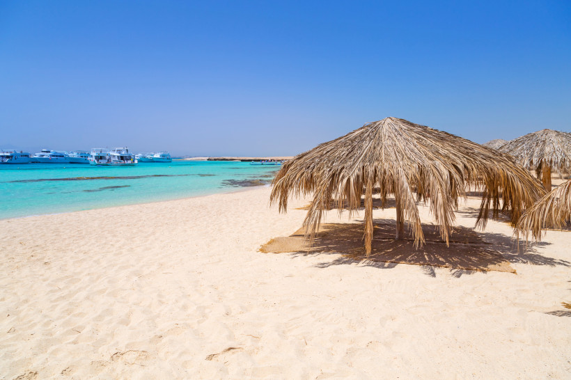 Strand mit türkisfarbenem Wasser und einfachen Palmen-Sonnenschirmen auf hellem Sand, Boote liegen im ruhigen Meer