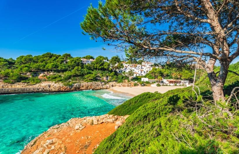 Mallorca - Cala Anguila Eine malerische, kleine Badebucht mit türkisfarbenem Wasser und feinem, hellen Sandstrand, umgeben von rötlichen Felsen und dichtem Pinienwald. Am Horizont sind einige weiße Häuser mit Terrakotta-Dächern zu sehen, die sich harmonisch in die grüne Landscha