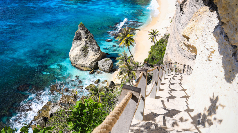 Nusa Penida, Bali Steile Treppe mit Seilgeländer führt an einer Felswand hinunter zu einem traumhaften Strand mit türkisblauem Wasser und Palmen