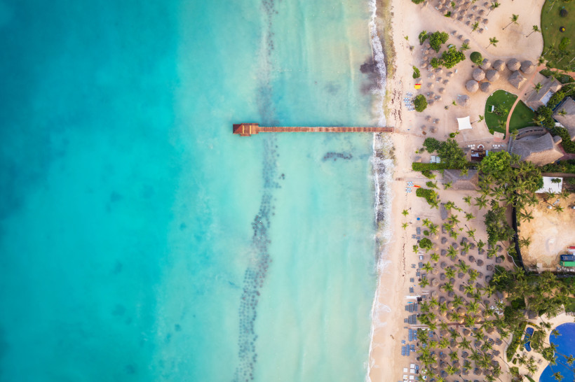 Drohnenaufnahme eines tropischen Strandes mit Holzsteg im türkisblauen Meer, Palmen und Liegestühlen entlang des Sandstrandes