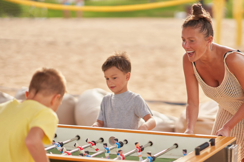 Ägypten - Hurghada Eine lachende Frau spielt mit zwei kleinen Jungen Tischfußball im Freien. Im Hintergrund ist ein Strand mit gelbem Volleyballnetz zu sehen. Die Szene wirkt sommerlich und entspannt