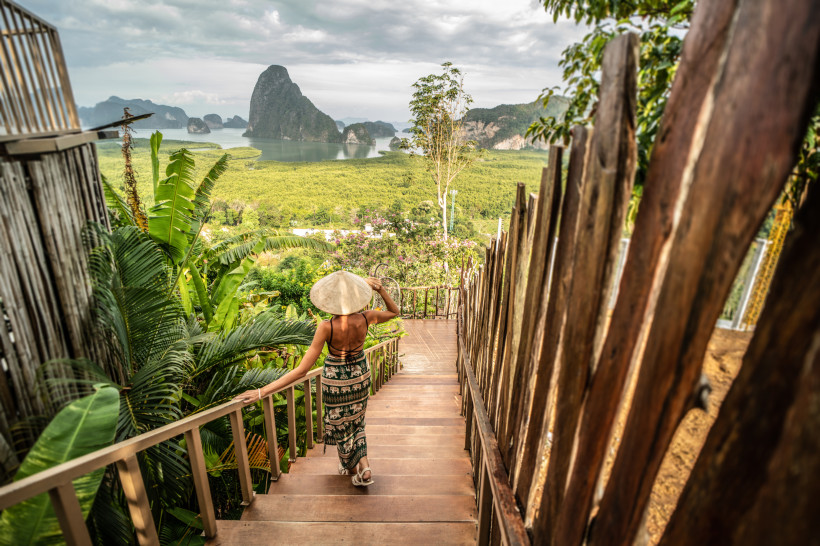Panoramablick über Phang Nga – Aussichtspunkt mit tropischer Natur in Thailand Frau mit Hut geht eine Holztreppe zum Aussichtspunkt hinunter und blickt auf die Kalksteinlandschaft und Mangroven von Phang Nga in Thailand.