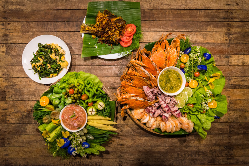Thailand - Krabi Das Bild zeigt eine bunte Auswahl traditioneller thailändischer Speisen, angerichtet auf einem rustikalen Holztisch. Rechts befindet sich eine große Platte mit gekochten Meeresfrüchten – darunter Garnelen, Tintenfisch und Krabben – garniert mit Limetten,