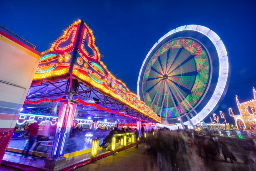 Mallorca Bunte Kirmesszene bei Nacht mit einem leuchtenden Riesenrad im Hintergrund und einem hell erleuchteten Autoscooter im Vordergrund. Viele Besucher schlendern durch das Gelände. Die bunten Lichter erzeugen Bewegungsspuren, die die dynamische Atmosphäre des
