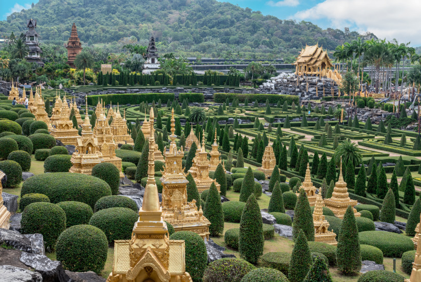 Prächtige goldene Tempel und geometrische Gärten in Nong Nooch Tropical Garden Goldene Tempelstrukturen und kunstvoll geschnittene Büsche im Nong Nooch Tropical Garden in Thailand vor bewaldeten Hügeln