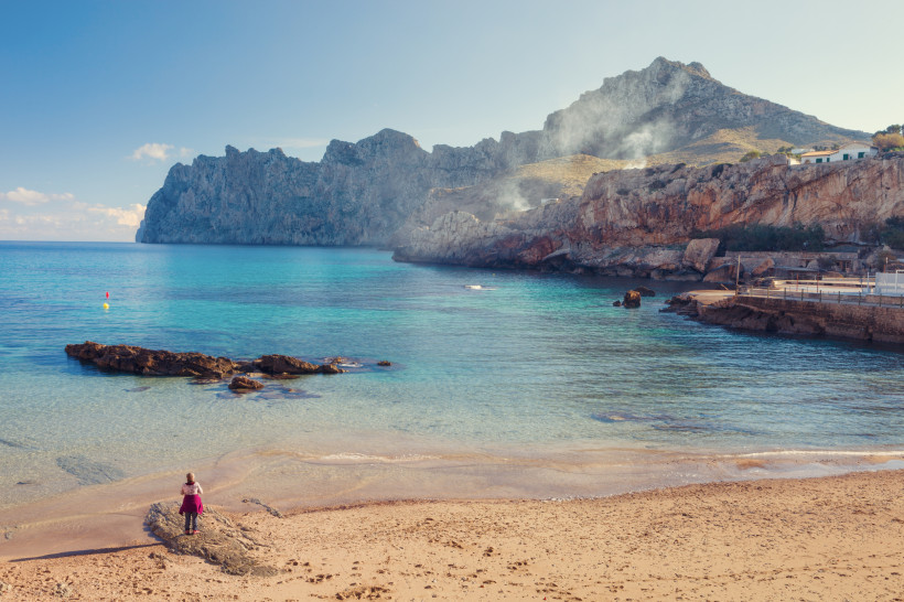 Cala Barques in Cala San Vicente mit Sandstrand, ruhigem Wasser und markanter Felskulisse der Serra de Tramuntana im Hintergrund.