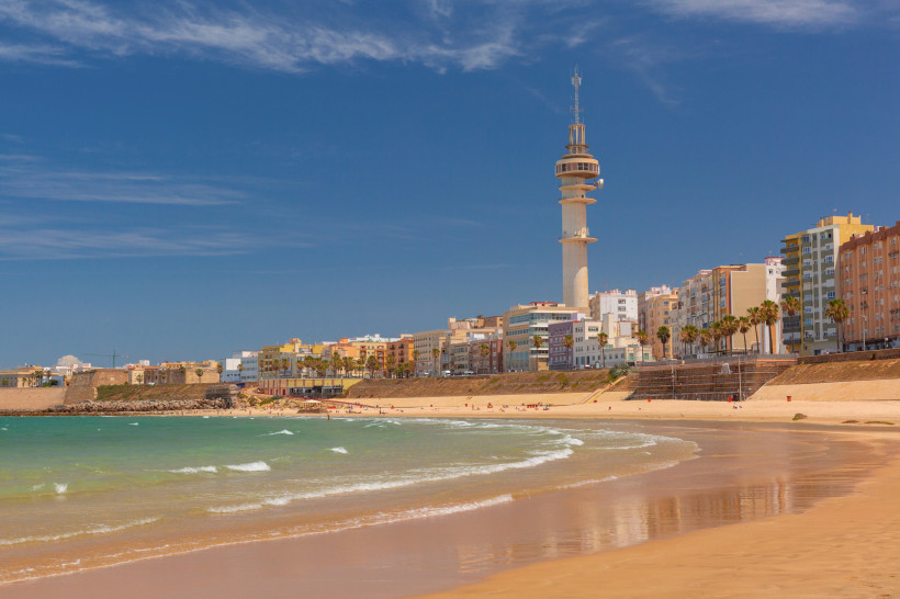 Blick auf den Strand La Caleta in Cádiz mit Küstenlinie und Turm Strand La Caleta mit Wellen, Promenade und markantem Turm vor der Skyline von Cádiz