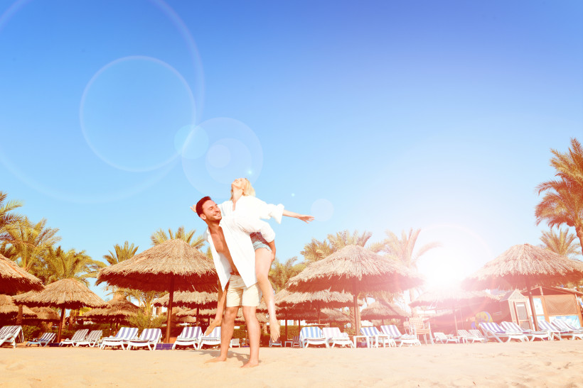 Ägypten Fröhliches Paar am Strand – der Mann trägt die Frau auf dem Rücken. Beide lachen, im Hintergrund sind Strohdachschirme, Palmen und leere Sonnenliegen unter einem strahlend blauen Himmel mit Sonnenreflexionen.