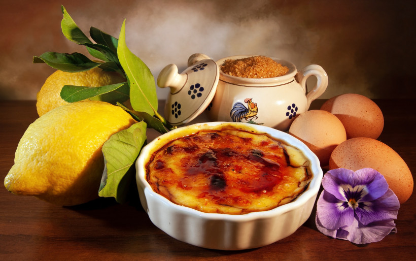 Crema Catalana in einer Keramikform mit karamellisierter Zuckerkruste, daneben Zitronen, Eier und ein Gefäß mit Zucker