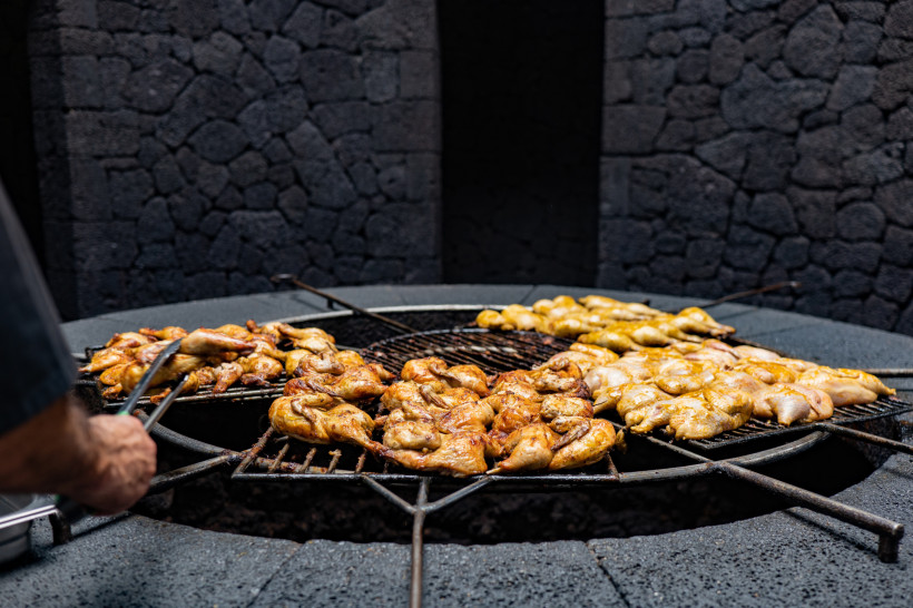 Traditionelles Kochen in Mancha Blanca – Grillen über Lavastein auf Lanzarote Traditionelles Grillen über Lavastein in Mancha Blanca auf Lanzarote, bei dem Fleisch auf einem runden Stein-Grill zubereitet wird.