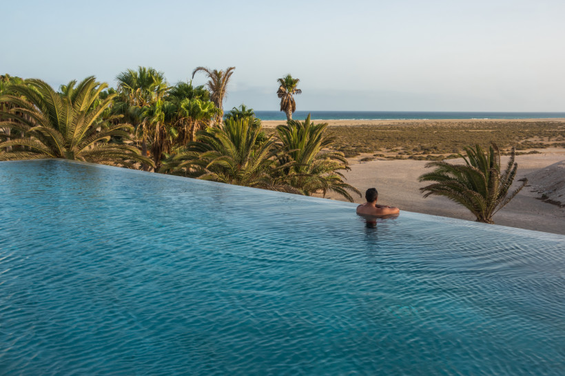 Fuerteventura Ein Mann lehnt entspannt am Rand eines Infinity-Pools und blickt auf Palmen, eine weite Dünenlandschaft und das Meer in der Ferne