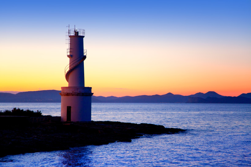 Formentera - La Savina Weißer Leuchtturm am Meer mit spiralförmiger Außentreppe, aufgenommen bei Sonnenuntergang. Der Himmel leuchtet in kräftigen Orangetönen, im Hintergrund sind die Umrisse von Bergen zu sehen.