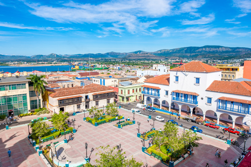 Blick auf den zentralen Parque Céspedes in Santiago de Cuba mit dem Ayuntamiento, kolonialen Gebäuden, Palmen und dem Meer im Hintergrund – beliebter Treffpunkt im Stadtzentrum.
