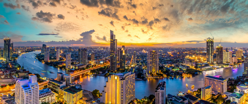 Bangkok Atemberaubende Panoramaaufnahme der Skyline von Bangkok bei Sonnenuntergang. Der Fluss Chao Phraya schlängelt sich durch die beleuchtete Stadt, gesäumt von modernen Hochhäusern und Brücken. Der Himmel leuchtet in dramatischen Orange-, Gelb- und Blautönen