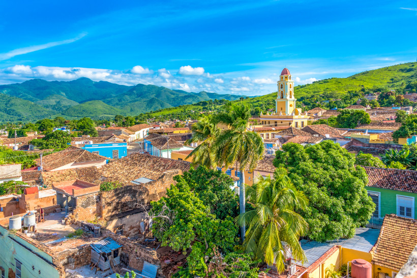 Panoramablick über die historische Altstadt von Trinidad auf Kuba mit farbenfrohen Kolonialhäusern, Palmen, dem gelben Glockenturm der Kirche und der Sierra del Escambray im Hintergrund.