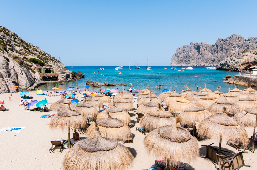 Cala Molins in Cala San Vicente mit Sandstrand, Sonnenschirmen und ruhigem, klarem Wasser vor der Felskulisse der Nordküste Mallorcas.