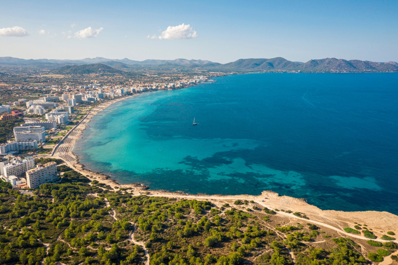 Mallorca - Cala Millor Eine ruhige Küstenlinie mit kristallklarem, türkisfarbenem Wasser, das sanft in den hellen Sandstrand übergeht. Viele Badegäste im Wasser und am Strand. Im Hintergrund eine grüne, leicht hügelige Landschaft. Der Himmel ist wolkenlos und blau.
