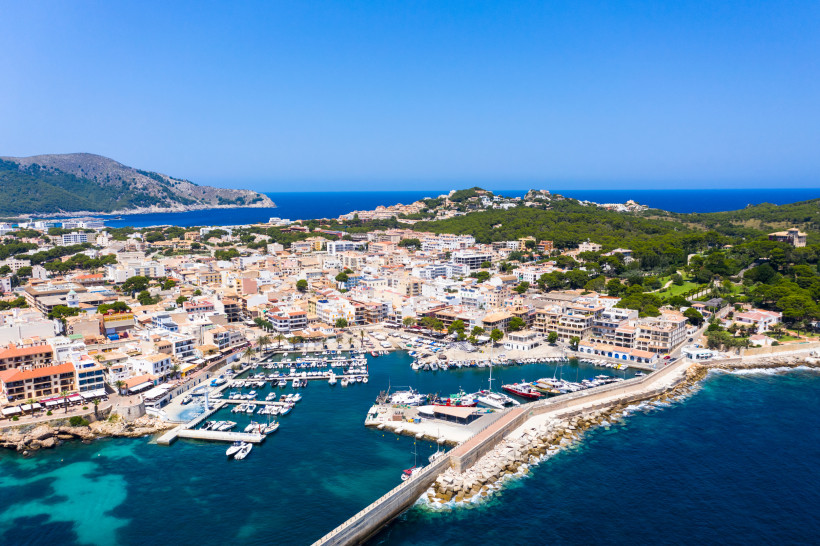 Mallorca - Cala Ratjada Luftaufnahme der Stadt Cala Ratjada mit Blick auf den Hafen, in dem zahlreiche Boote und Yachten liegen. Die Stadt ist dicht bebaut mit hellen, mediterranen Häusern, teils mit Flachdächern und Dachterrassen. Eine Promenade verläuft entlang des Wassers, ge