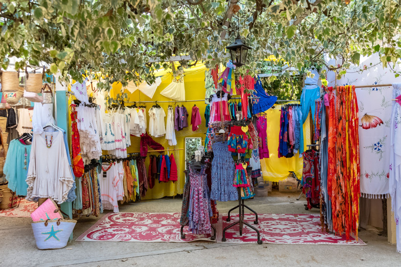 Ibiza - Hippiemarkt Punta Arabí Bunter Hippiemarkt auf Ibiza: Unter einem Blätterdach hängen und liegen farbenfrohe Kleidungsstücke wie Kleider, Tuniken und Tücher. An einem Kleiderständer sind Sommerkleider in kräftigen Farben ausgestellt. Die Marktstände sind mit gelben Stoffbahnen de